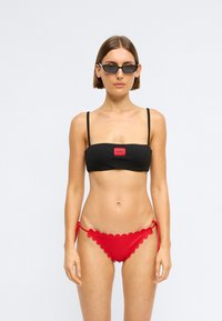 Mustmustriibuline must bikiinipealne, millel on spagetitripid ja punane logo silt; punased lainelised bikiinialused reguleeritavate sidemetega külgedel.