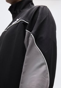 Μαύρο αθλητικό μπουφάν με γκρι λεπτομέρειες, που διαθέτει ψηλό γιακά και λευκή ραφή κατά μήκος των ραφών. Λείο ύφασμα με εφαρμοστό σχέδιο.