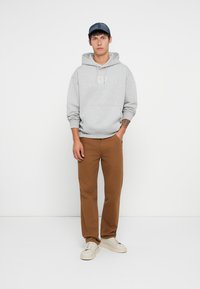 Manta cinza com logotipo, calças de moletom castanhas e ténis brancos. O modelo usa um boné azul. Conjunto simples e casual em frente a um fundo neutro.