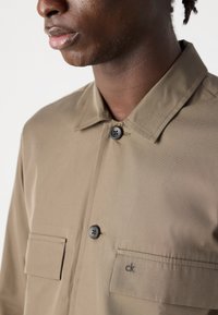 Khaki button-up shirt featuring a collar, two chest pockets, and black button details. Smooth fabric with an embroidered logo.