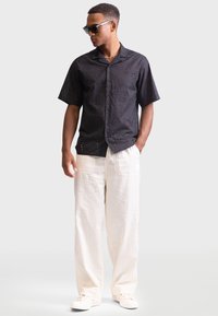 Black short-sleeve shirt with detailed patterns, paired with light beige wide-leg linen pants and white sneakers. Model wearing sunglasses.