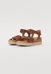 Brown leather sandals with crisscross straps, textured wedge sole, and a light-colored rubber base. Features cushioned insole for comfort.