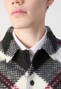 Wool-blend shirt jacket featuring a black, white, and red check pattern, a pointed collar, and black buttons. White shirt underneath visible.