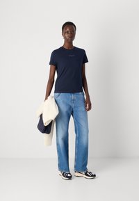 T-shirt blu marine a maniche corte, in cotone, abbinato a jeans a gamba dritta di colore azzurro chiaro. Tiene in mano un maglione color crema e blu scuro, indossa sneakers.