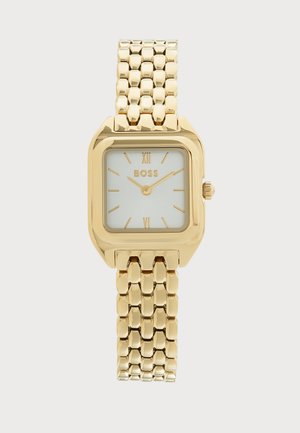 Reloj de pulsera dorado BOSS con esfera cuadrada de color claro, números romanos y una pulsera metálica de eslabones sobre un fondo claro.