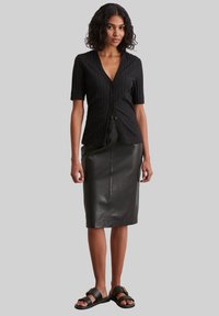 Schwarze, gerippte kurzärmelige Knopfhemd, kombiniert mit einem taillierten schwarzen Leder-Midirock und schwarzen flachen Sandalen. Glatte Texturen und taillierter Schnitt.