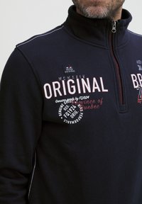 Felpa zip con colletto alto di colore blu navy, con testo e loghi stampati in bianco e rosso, e cuciture a contrasto lungo le cuciture.