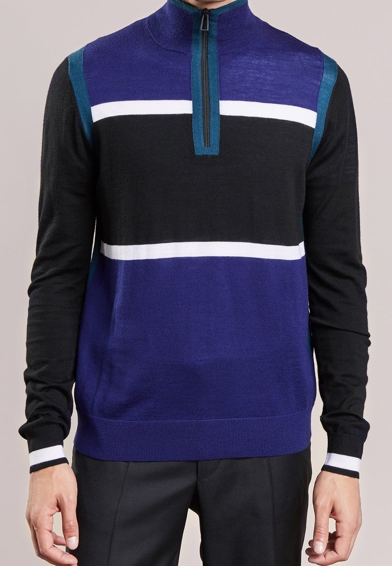 Homme portant un pull à manches longues avec des rayures horizontales bleu, noir, blanc et bleu sarcelle et une fermeture éclair partielle au col.