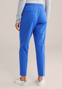 Plave uske hlače od teksturiranog materijala s glatkim završetkom, s dvije stražnje džepa i elastičnim pojasom. Vidljive su bijele tenisice.