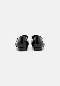 Mocasines de charol negro con parte trasera redondeada, que presentan un detalle de lazo y tacones bajos en bloque, con una textura suave y un acabado brillante.