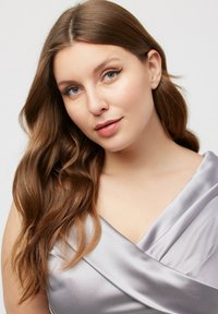 Stříbrné saténové šaty s překříženým materiálem na výstřihu, hladká textura a jemný lesk. Modelka má dlouhé, vlnité hnědé vlasy a přirozený make-up.