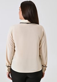 Frau mit langen dunklen Haaren, die eine beigefarbene Bluse mit langen Ärmeln und schwarzem Besatz am Kragen und an den Manschetten trägt, stehen vor einem schlichten Hintergrund.