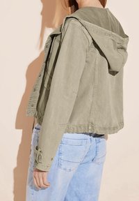 Personne portant une veste à capuche vert olive clair et un jean bleu clair, vue de dos sur un fond beige.