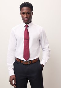 White dress shirt with a pointed collar, paired with a maroon textured tie featuring a silver tie bar. Dark trousers and a brown leather belt complete the look.