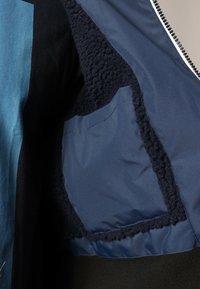 Veste colorée composée de panneaux bleus, noirs et navy, avec une texture douce et pelucheuse autour du col et une poche à rabat.