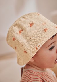 Chapeau seau jaune avec motifs de soleil, tissu texturé, accents cousus et une étiquette comportant une icône de soleil.