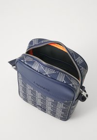Borsa a tracolla Lacoste in blu navy con un motivo geometrico. Caratterizzata da un interno arancione a contrasto, chiusura con zip e una superficie esterna sintetica liscia.