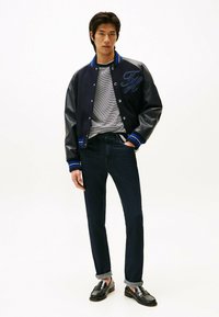 Zwarte varsityjack met blauwe strepen en letters, grijs gestreept t-shirt, donkere denim jeans en zwarte instappers. Gladde, casual stijl.