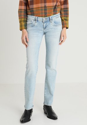 Persona con vaqueros azul claro, botines negros y camisa de manga larga de cuadros en naranja, verde y marrón, de pie frente a un fondo liso.