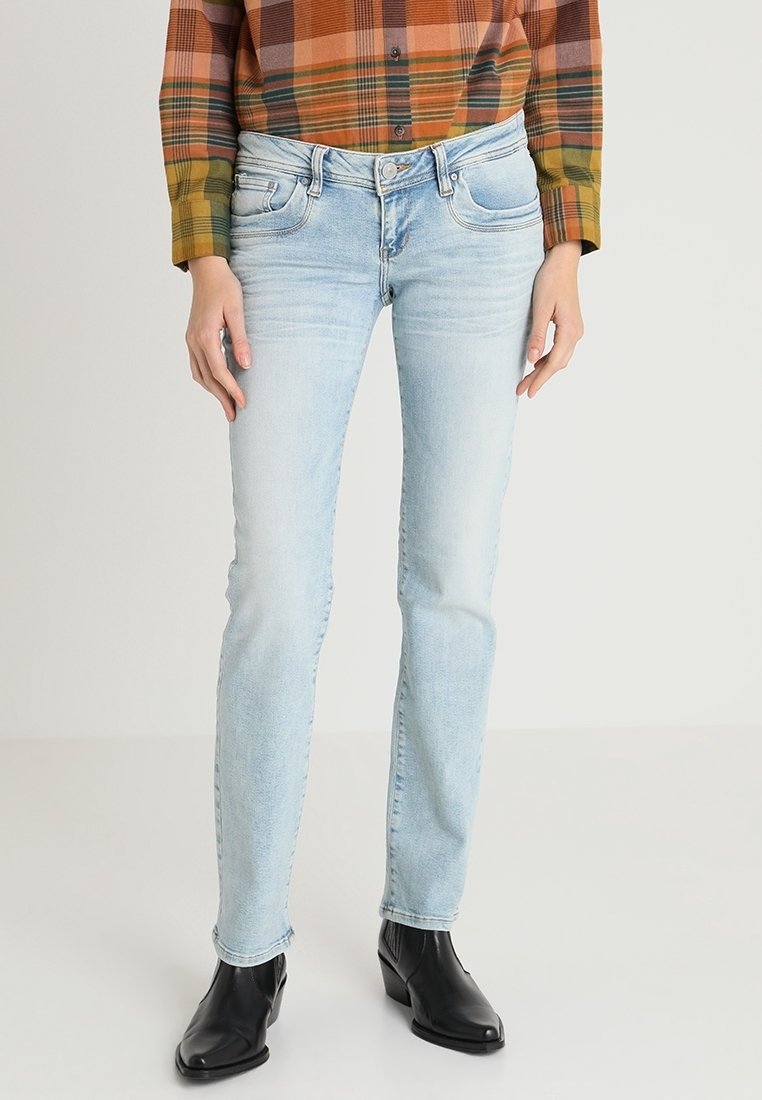 Personne portant un jean bleu clair, des bottines noires et une chemise à carreaux orange, verte et brune à manches longues, debout sur un fond uni.
