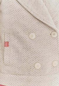 Dvojité sako v světle šedé barvě s herringbone vzorem, obsahující bílé knoflíky, boční kapsu a malou růžovou nášivku.