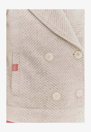 Dwurzędowy płaszcz w jasnoszarym materiale w jodełkę, z białymi guzikami, boczną kieszenią i małym różowym akcentem w postaci metki.