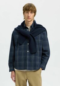 Camicia a quadri blu navy con dettagli bianchi, fronte con bottoni, abbinata a un maglione blu navy scuro annodato intorno alle spalle. Pantaloni beige completano l'outfit.