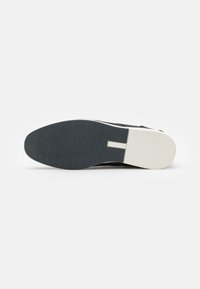 Semelle de chaussure noire avec une surface texturée ; présente un bord blanc et une rainure centrale pour plus de flexibilité ; fabriquée en caoutchouc et en matériaux synthétiques.