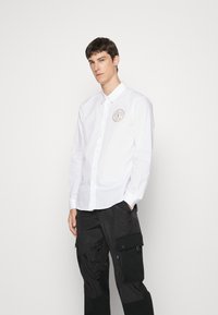 Camicia bianca a maniche lunghe con colletto button-down, dotata di un patch circolare con logo sul petto. Indossata con pantaloni cargo neri.