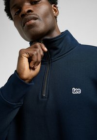 Pull bleu marine avec un col zippé, fabriqué en tissu texturé. Présente un logo "Lee" blanc sur la poitrine.