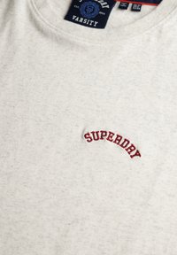 Jasnoszara bawełniana koszulka z okrągłym dekoltem, ozdobiona czerwonym, wyhaftowanym logo "SUPEDRAY" na piersi oraz żebrowanym kołnierzykiem.