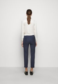 Biała, przejrzysta bluzka z długim rękawem i plisami na ramionach, zestawiona z granatowymi, zwężanymi spodniami z tylnymi kieszeniami, noszona z czarnymi butami.