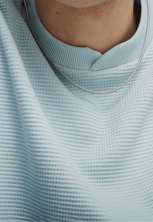 Detailní záběr na krk, který nosí tenký stříbrný řetízek přes svetřík s jemnou modrou texturou ve stylu vaflí.