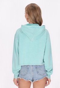 Sudadera corta en tejido de color aqua claro que cuenta con capucha con cordones y puños acanalados, combinada con pantalones cortos de denim deshilachados.