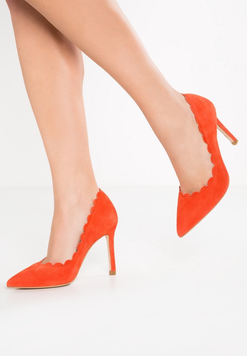 Des jambes portant des escarpins en daim orange vif avec des bords festonnés, posées sur un fond blanc uni.