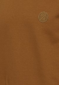 Tissu marron avec un petit logo rond brodé présentant les lettres C et F séparées par une ligne diagonale près du coin supérieur droit.