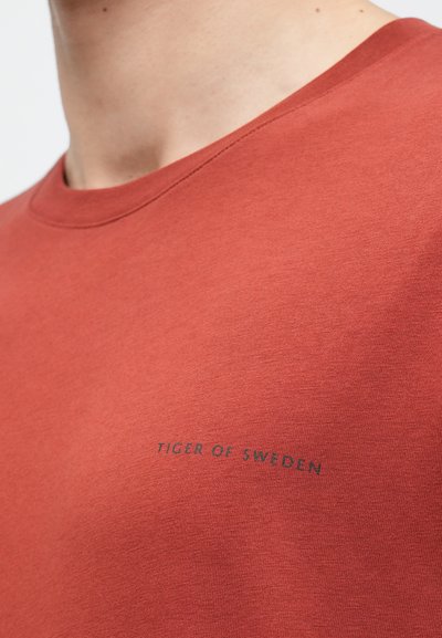 Ruda spalva medvilnės marškinėliai su apvalia iškirpte, priekyje esantis minimalistinis logotipas "TIGER OF SWEDEN" atspausdintas tamsiai mėlyna spalva.