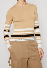 Pull long à manches longues beige en tricot avec des rayures horizontales blanches, noires et marron. Présente une texture côtelée et un logo en bas.