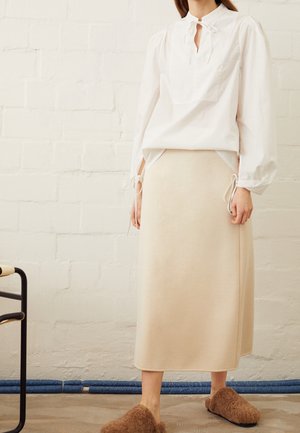 Femme portant un chemisier blanc à manches longues avec des détails de nouage, une jupe midi beige et des pantoufles marron pelucheuses, debout contre un mur en briques blanches.