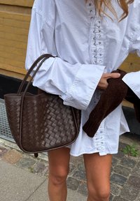 Borsa a tracolla in pelle intrecciata marrone con manici doppi e un design spazioso. Abbinata a una camicia bianca con dettagli allacciati e un maglione marrone.