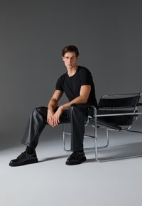 Crna majica, crne kožne hlače i crne cipele sa zakovicama. Model sjedi na elegantnoj kromiranoj stolici na pozadini u nijansama sive.