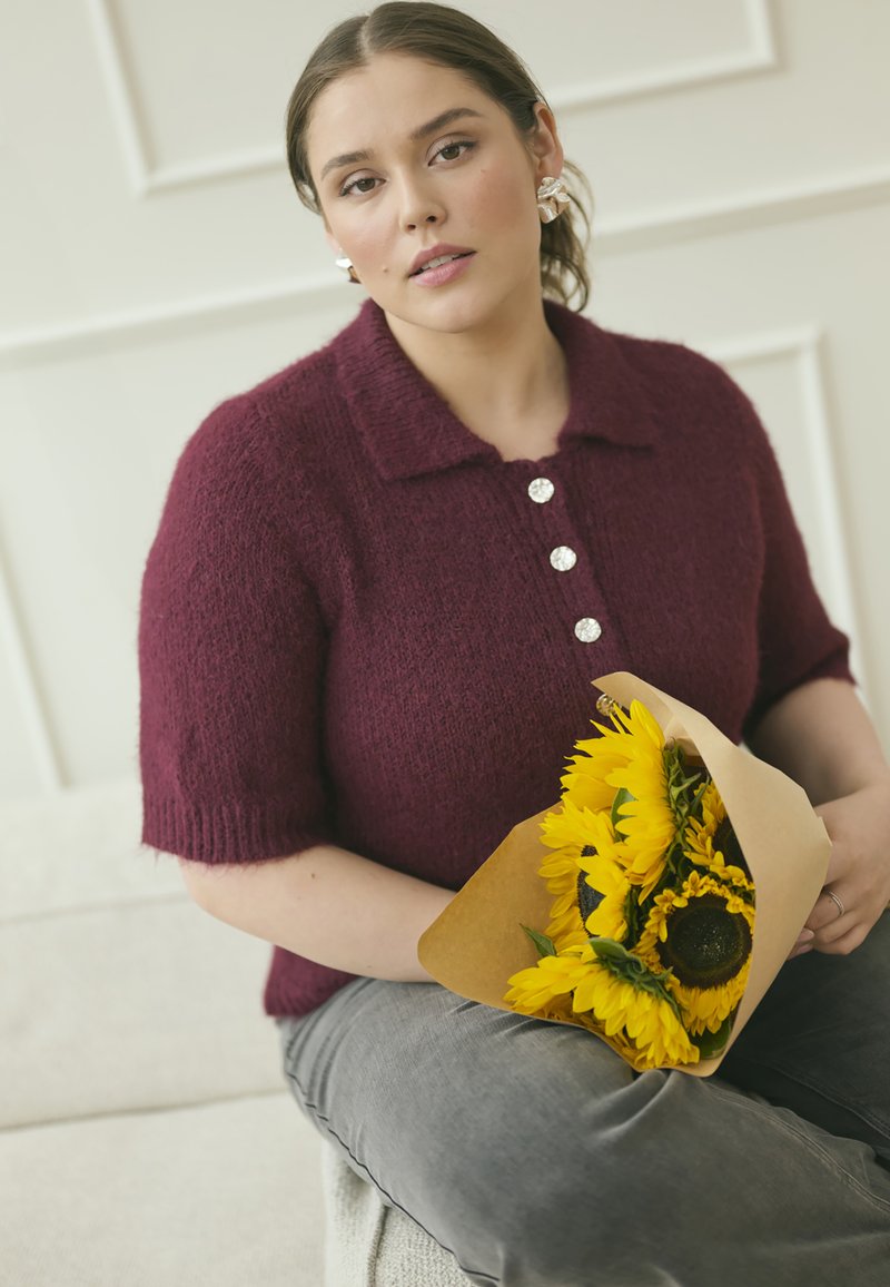 Pull en tricot bourgogne à manches courtes avec un col et cinq boutons. La personne tient un bouquet de tournesols jaunes enveloppé dans du papier.