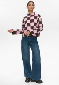 Maglione a maglia a motivo a quadri rosa e marrone con colletto alto, abbinato a jeans in denim blu a gamba larga e stivali neri chunky.
