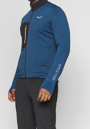 Man draagt een blauw jack met ritssluiting en een zwart borstzakje, met de tekst "SALEWA" op de mouw, gecombineerd met een zwarte broek.