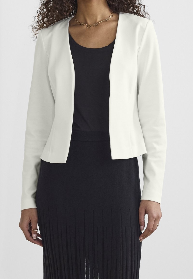 Blazer blanc raccourci avec une texture lisse et un devant ouvert, porté sur un haut noir et une jupe avec des détails à franges verticales.