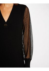 Robe noire avec un col en V, un corps en maille côtelée, et des manches bouffantes en dentelle transparente avec un motif floral. Finition avec un détail de bouton décoratif.