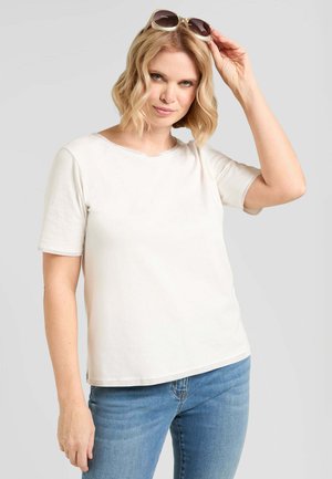 Donna bionda con maglietta bianca e jeans blu che solleva degli occhiali da sole marroni dalla testa, guardando verso la fotocamera.