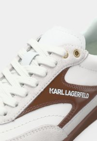 Hvit atletisk joggesko med brune detaljer, laget av semsket skinn og syntetiske materialer, runde lisser og en logo på siden. Gullfargede metallhull.