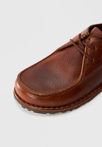 Brown leather lace-up shoe with textured finish and stitched detailing, featuring a rubber sole with a tread pattern.