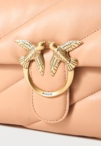 Saco de couro bege com um fecho dourado que apresenta duas aves gravadas a olhar-se uma para a outra e um anel rotulado "PINKO."